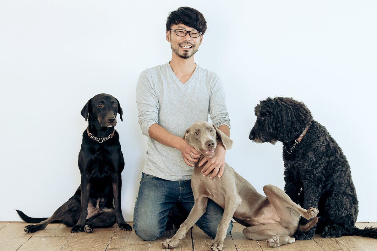 白壁の前に座る三頭の犬と、中央で膝をついたトレーナー